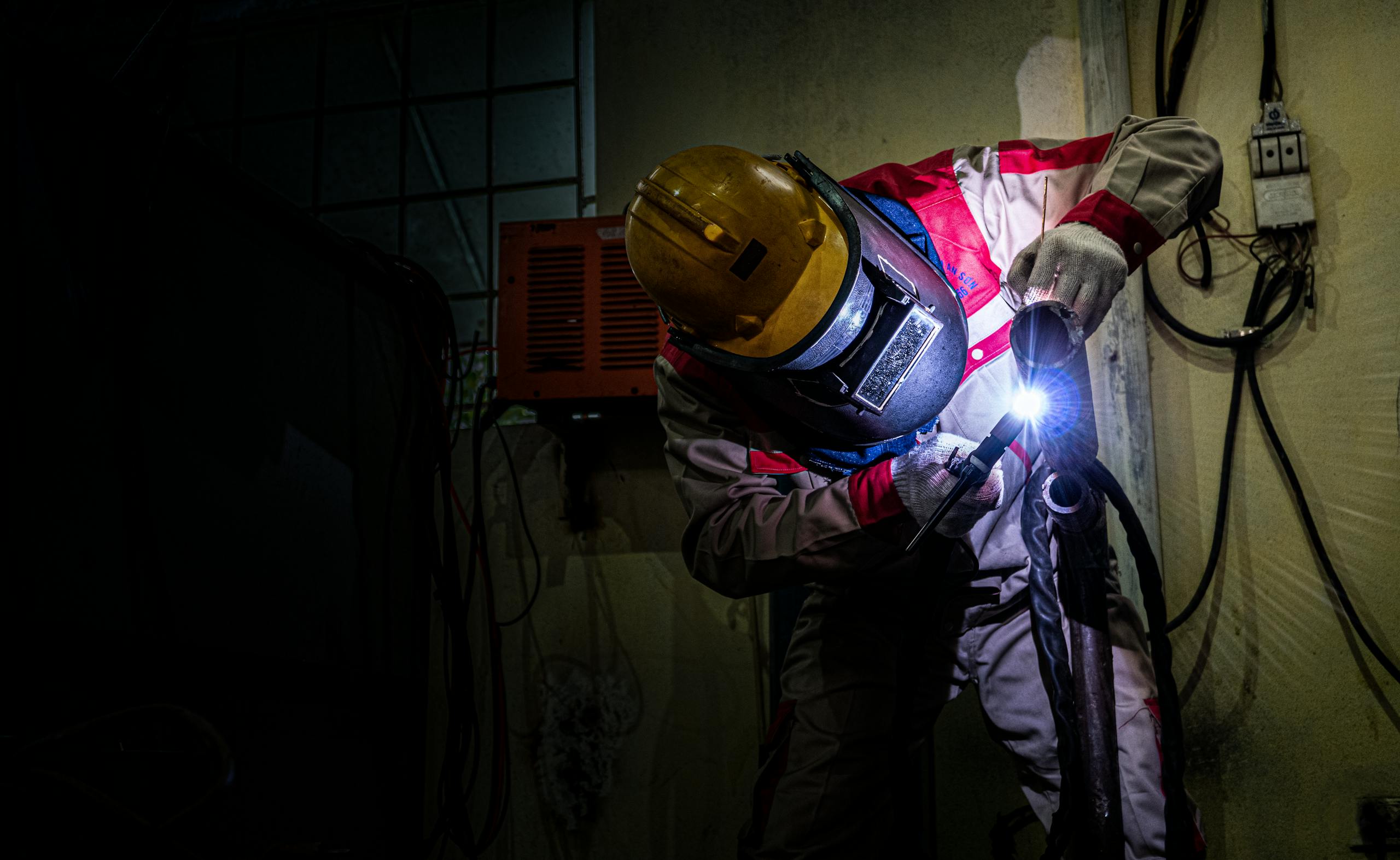 A welder wearing protective gear works with a torch indoors, highlighting industrial labor.